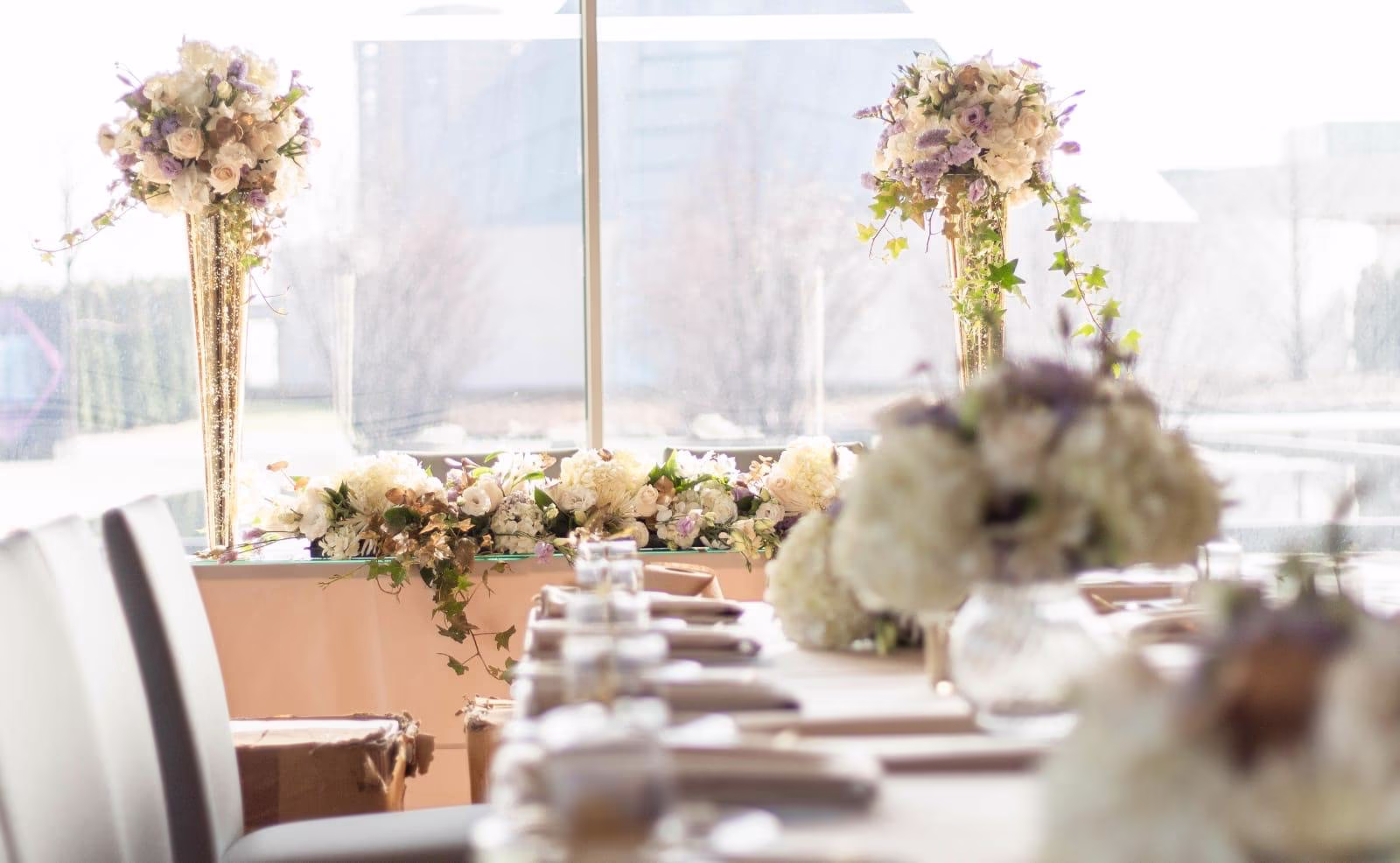 Reception head table with tall gold trumpet vase centrepieces of cream roses, lavender and white hydrangea