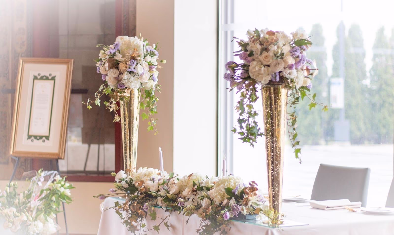 Tall gold trumpet vases of cream roses and lavender beside a framed seating sign at a reception