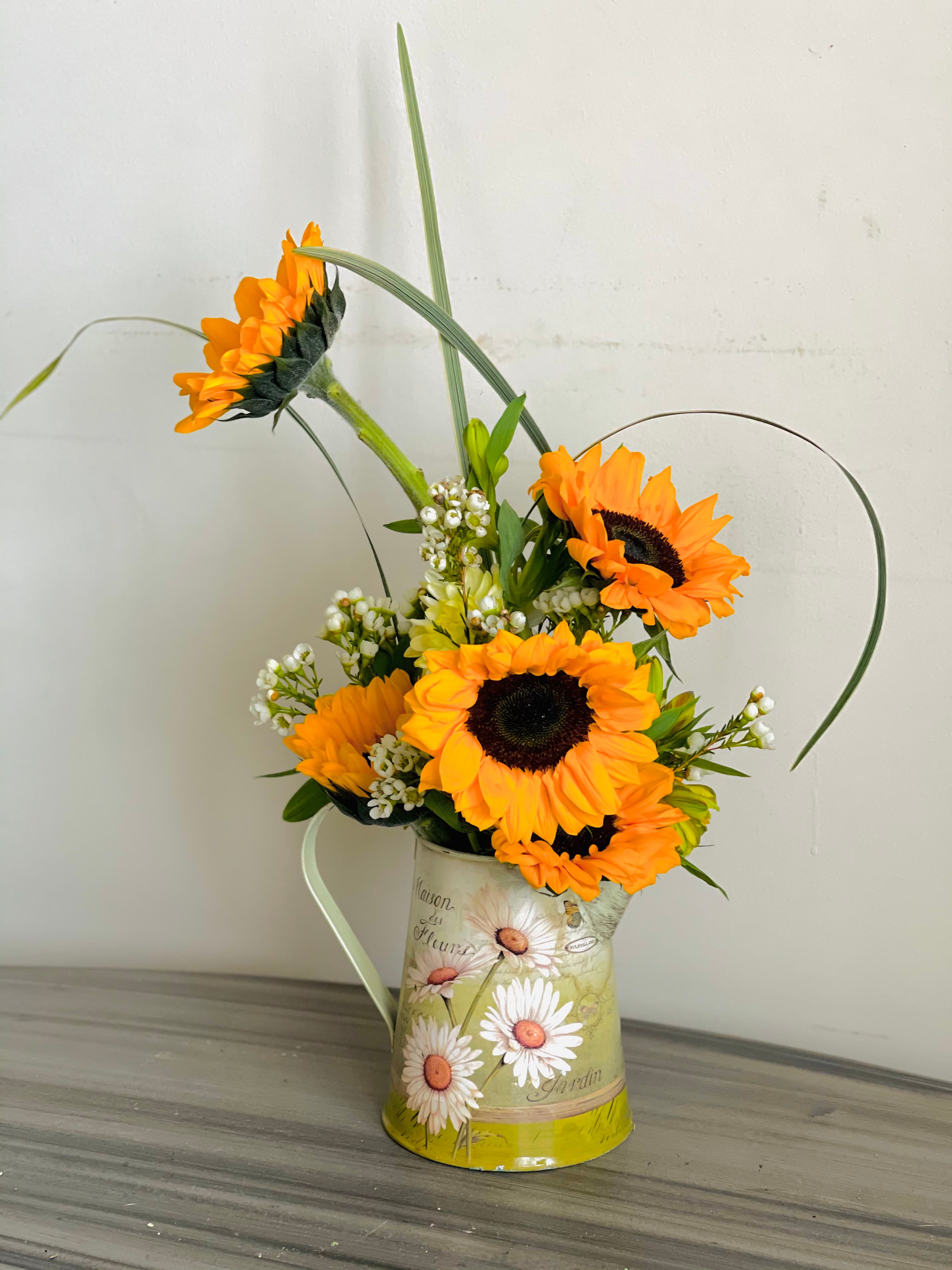 Filled with Sun arrangement of sunflowers, white waxflower, and looped grasses in a daisy-painted metal pitcher