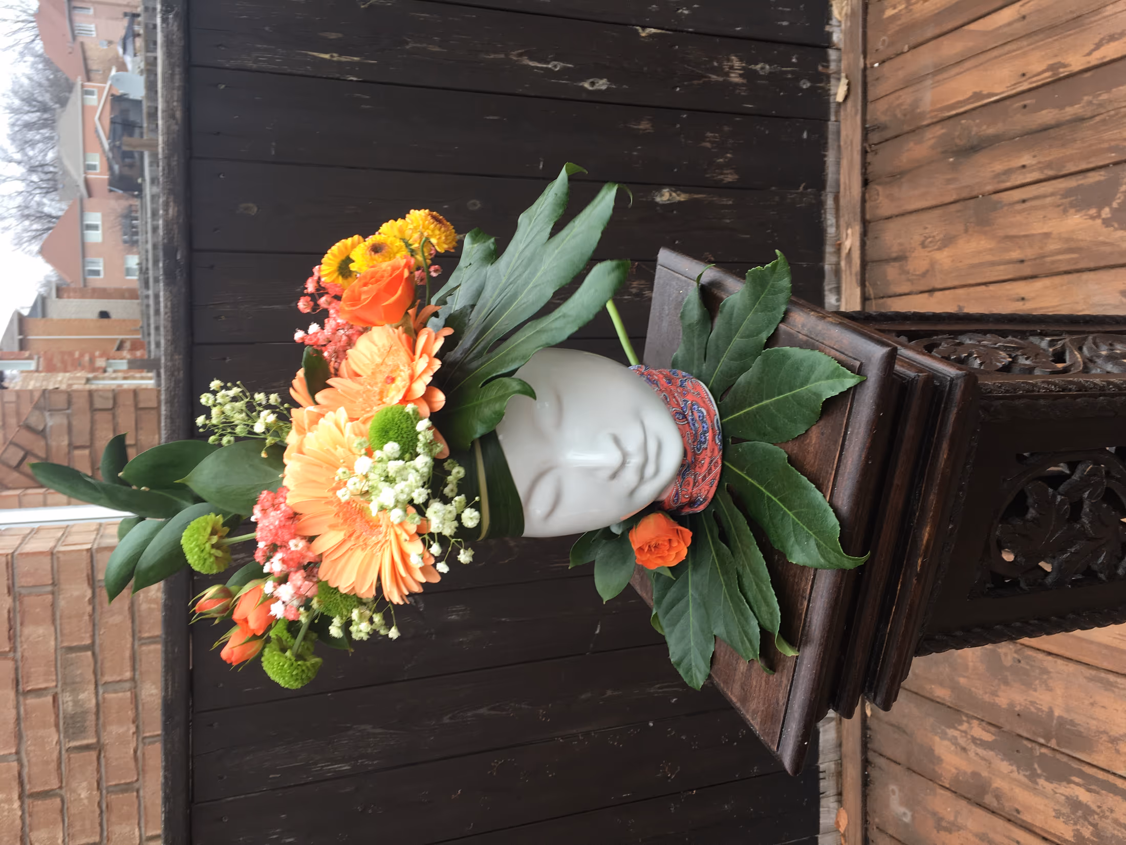 White ceramic face vase holding orange gerbera daisies, ranunculus, and lush greenery