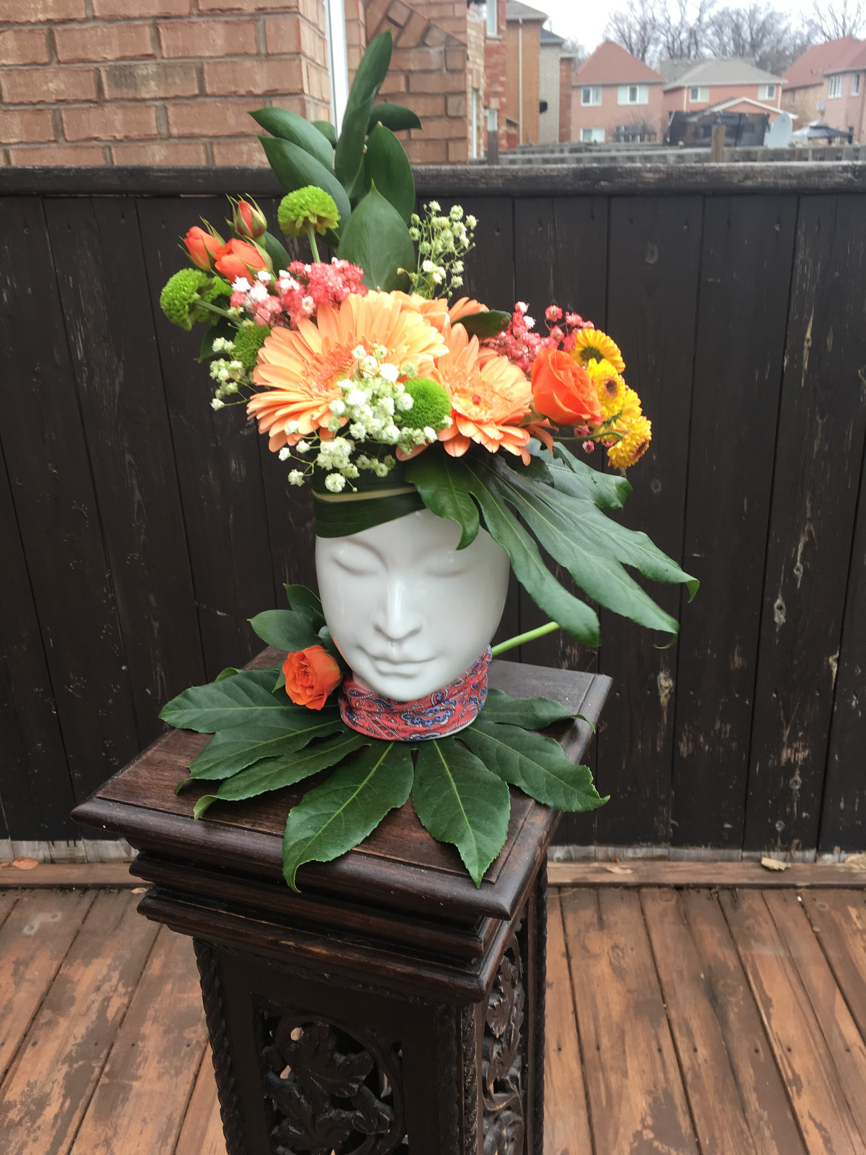 White ceramic face vase holding orange gerbera daisies, ranunculus, and lush greenery