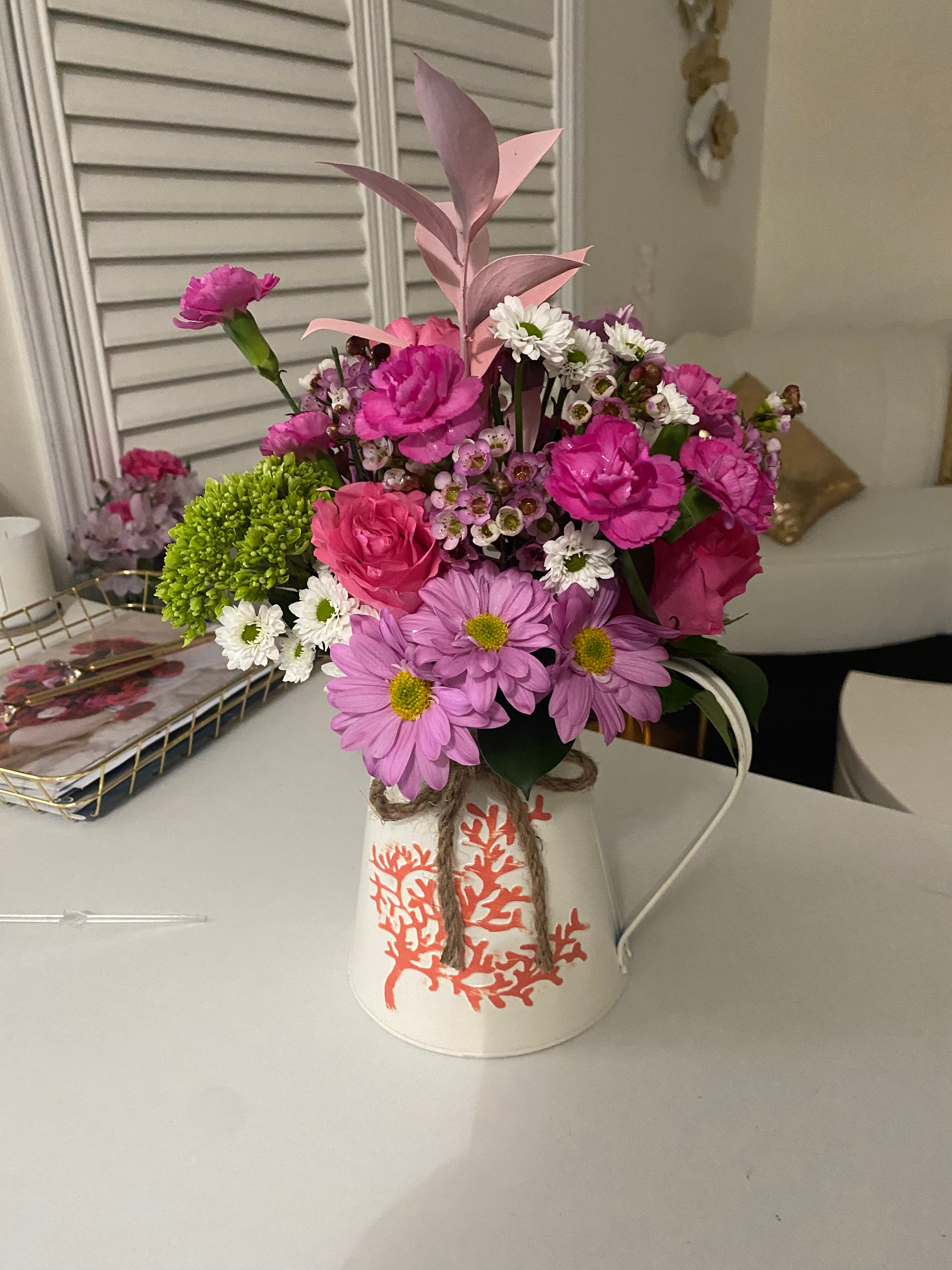 Pitcher of Expression arrangement with pink carnations, roses, daisies, and green blooms in a white pitcher painted with a red coral motif