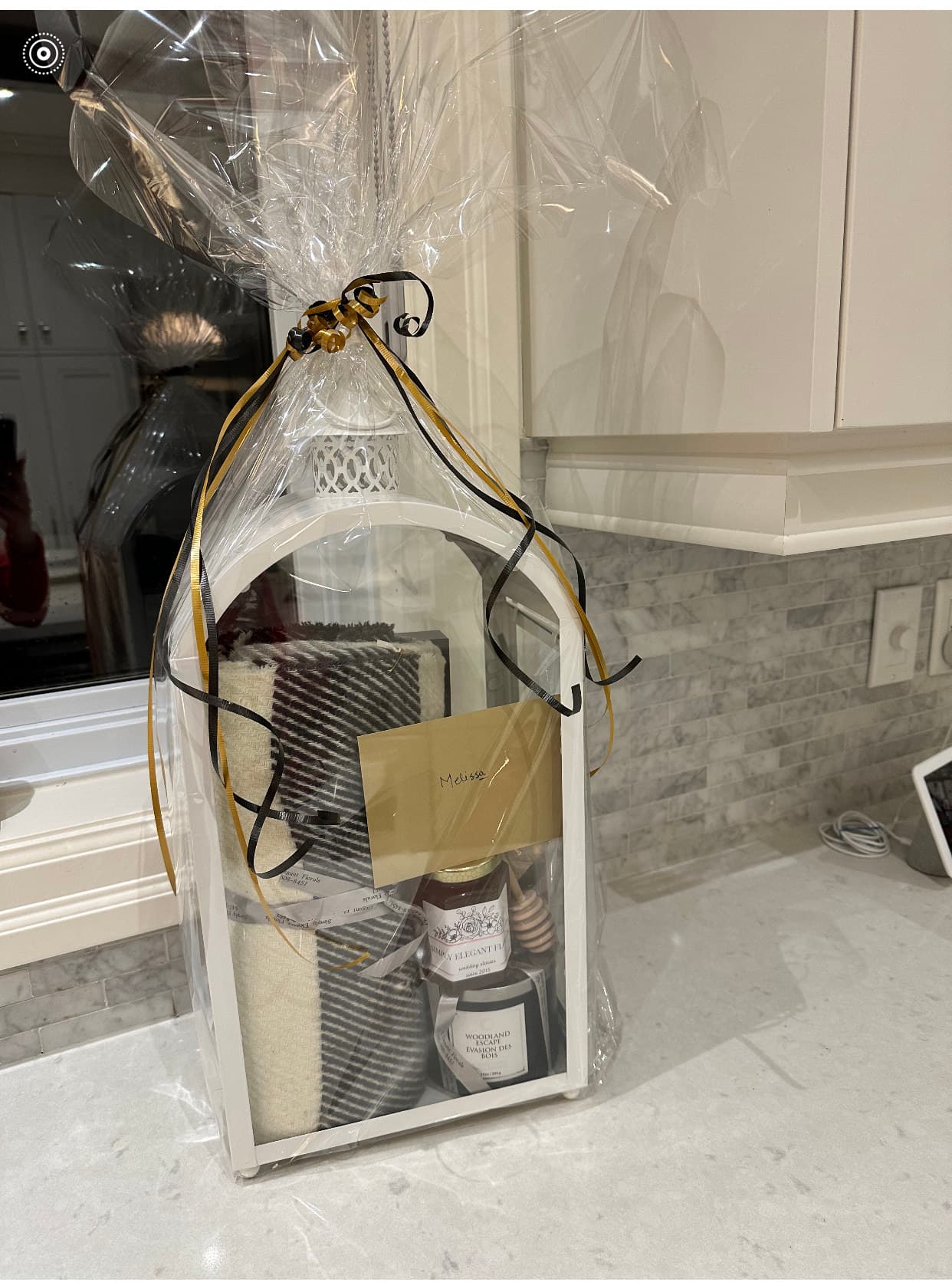 White lantern-style gift box with a folded throw, honey jar, and candle wrapped in cellophane with ribbon