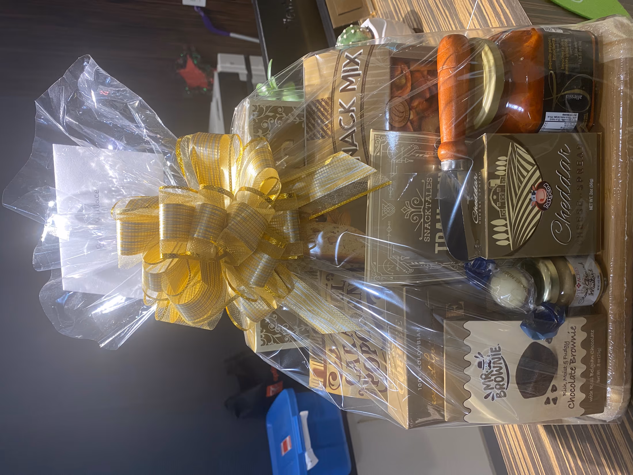 Gourmet gift hamper of snacks, chocolates, and preserves on a board, wrapped in cellophane with a gold bow