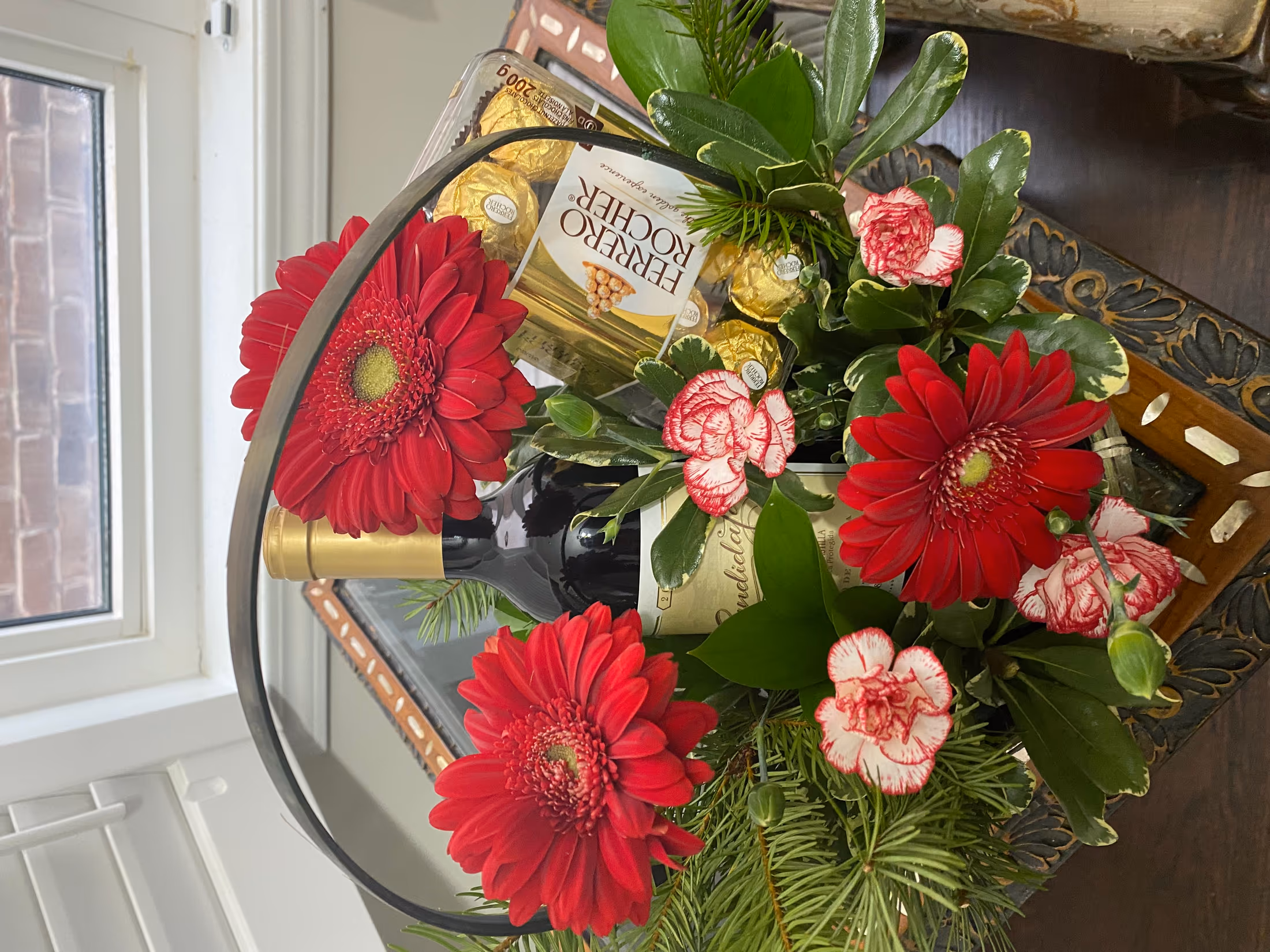 Willow basket with a wine bottle, Ferrero Rocher chocolates, red gerbera daisies, and striped carnations