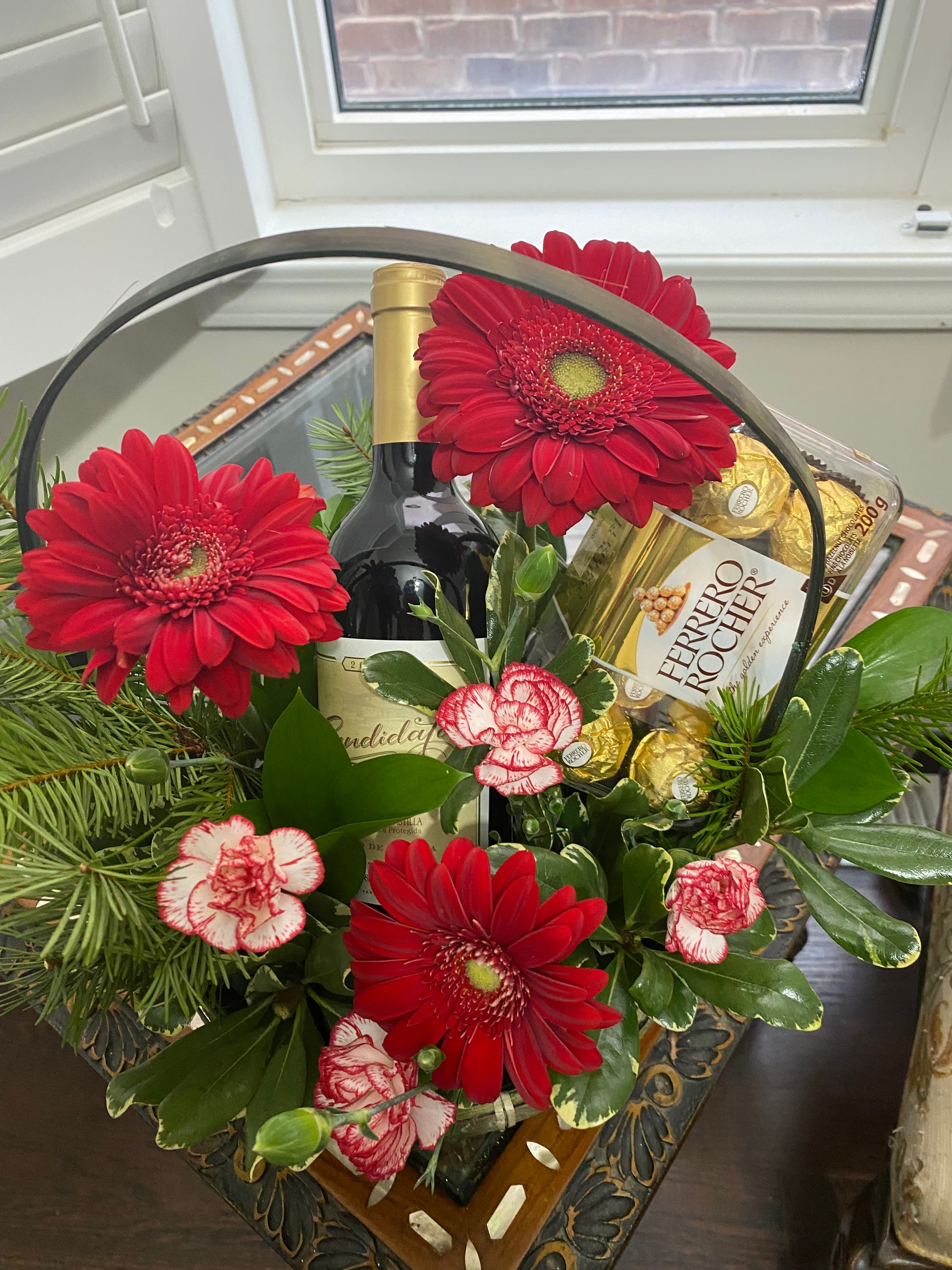 Willow basket with a wine bottle, Ferrero Rocher chocolates, red gerbera daisies, and striped carnations