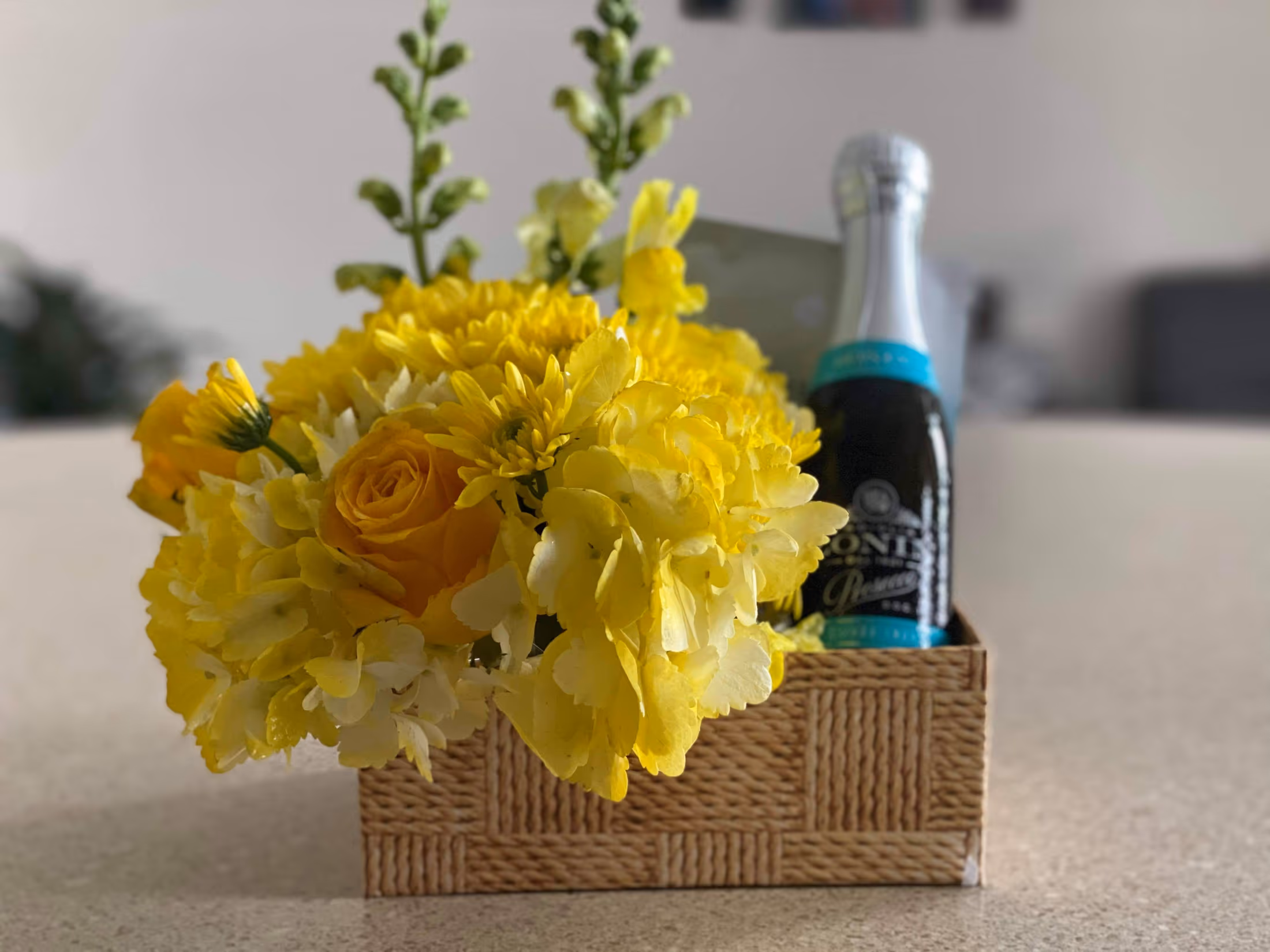 Woven-look box of yellow hydrangea, roses, and mums beside a sparkling wine bottle