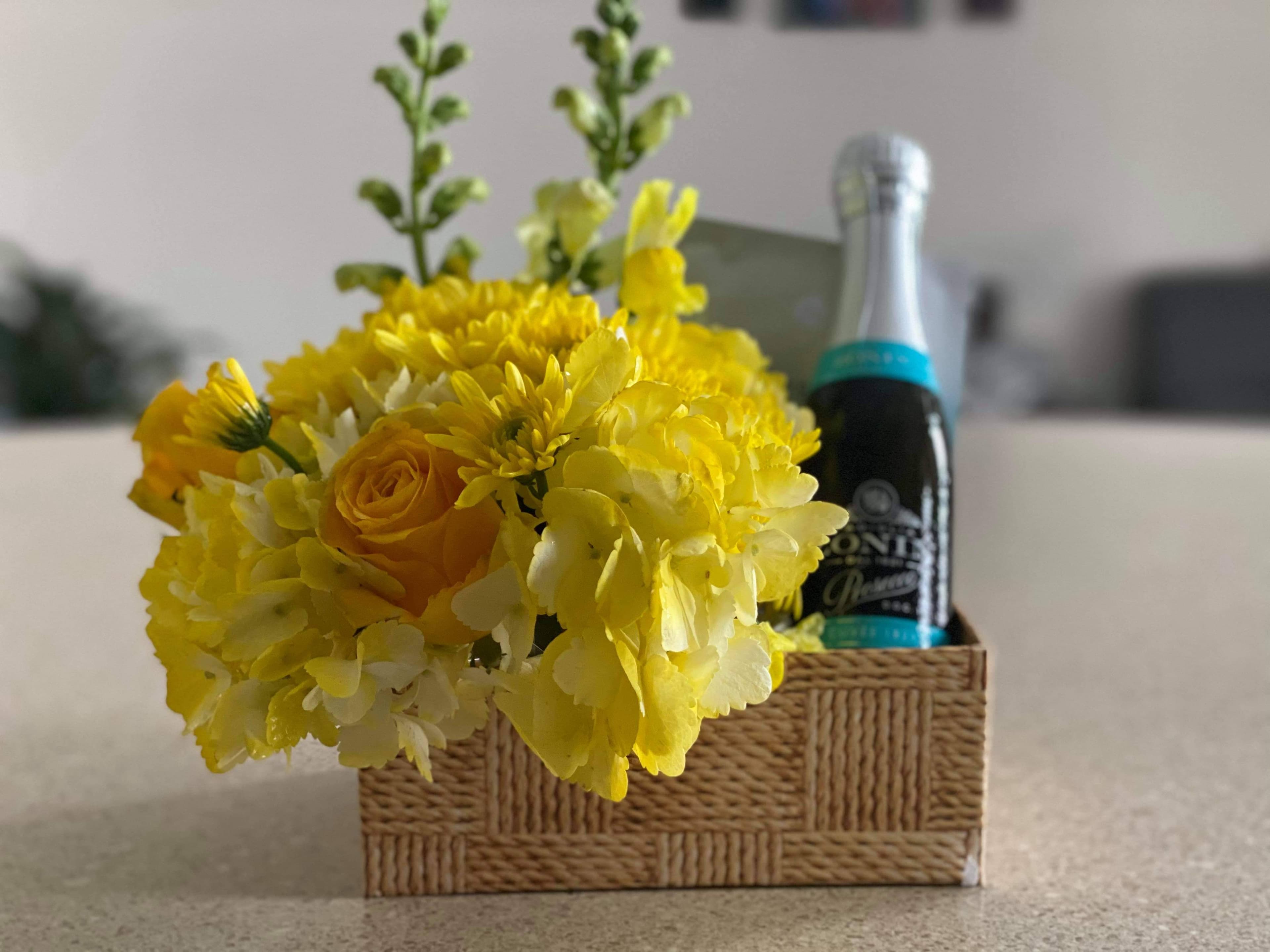 Woven-look box of yellow hydrangea, roses, and mums beside a sparkling wine bottle