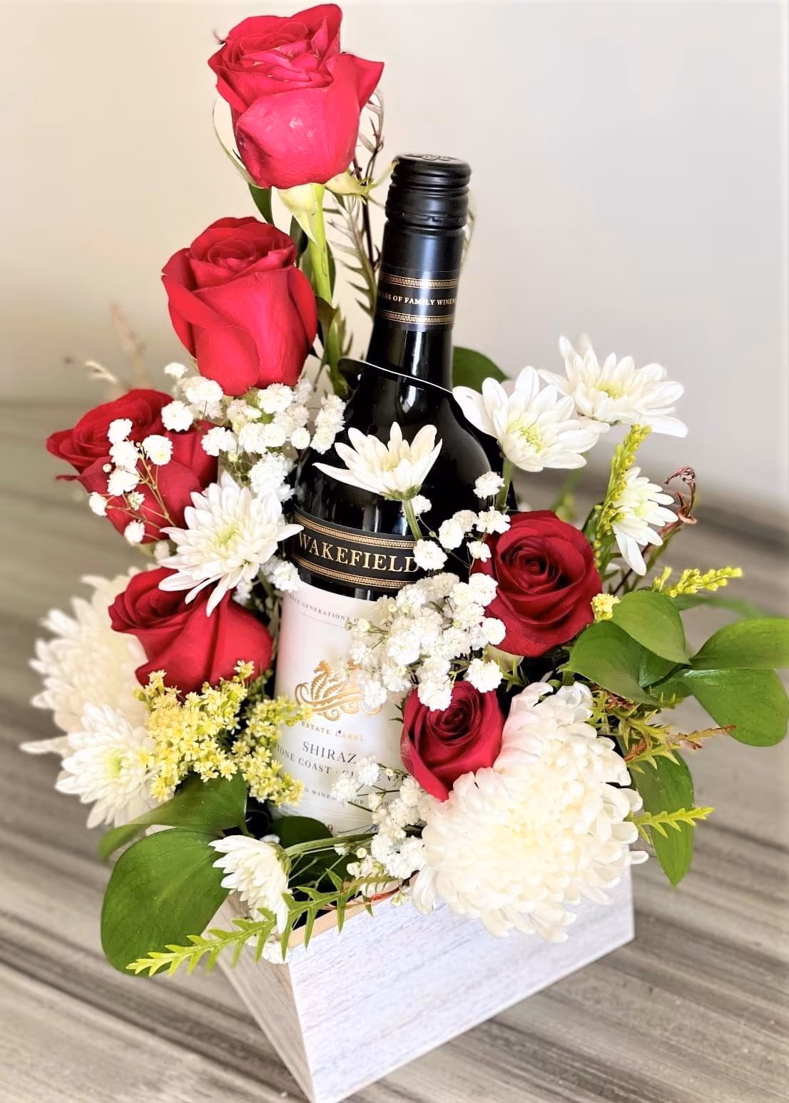 Whitewashed wooden box with a wine bottle surrounded by red roses, white chrysanthemums, and baby's breath