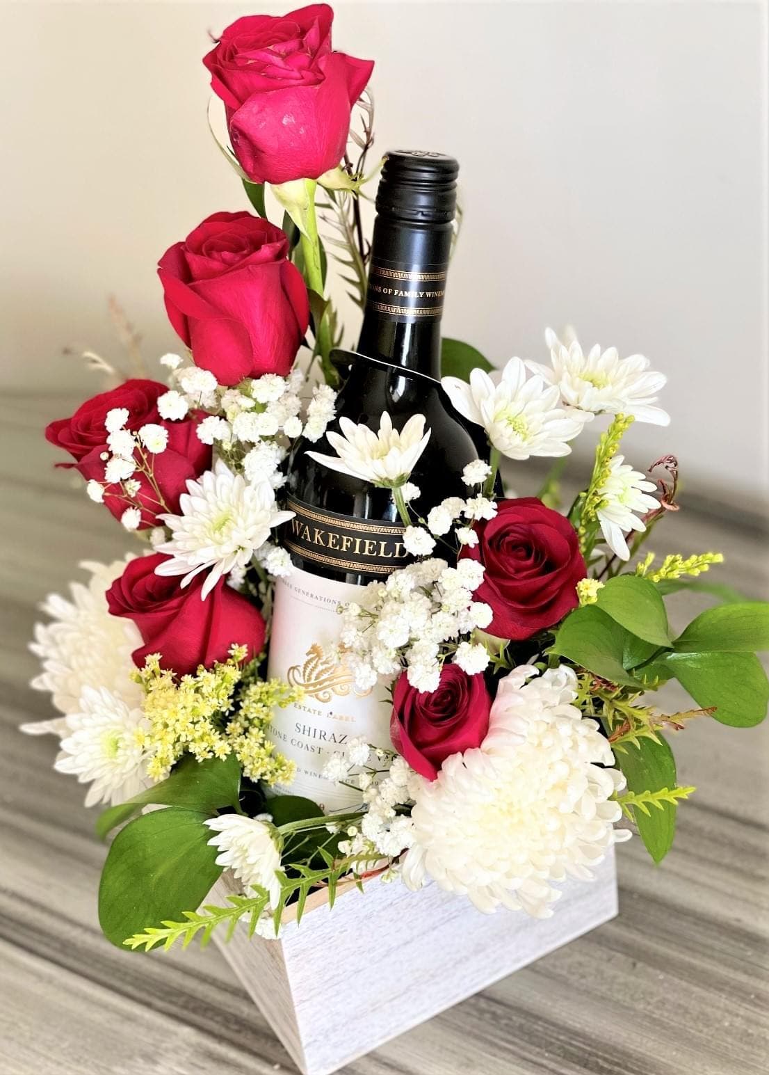 Whitewashed wooden box with a wine bottle surrounded by red roses, white chrysanthemums, and baby's breath