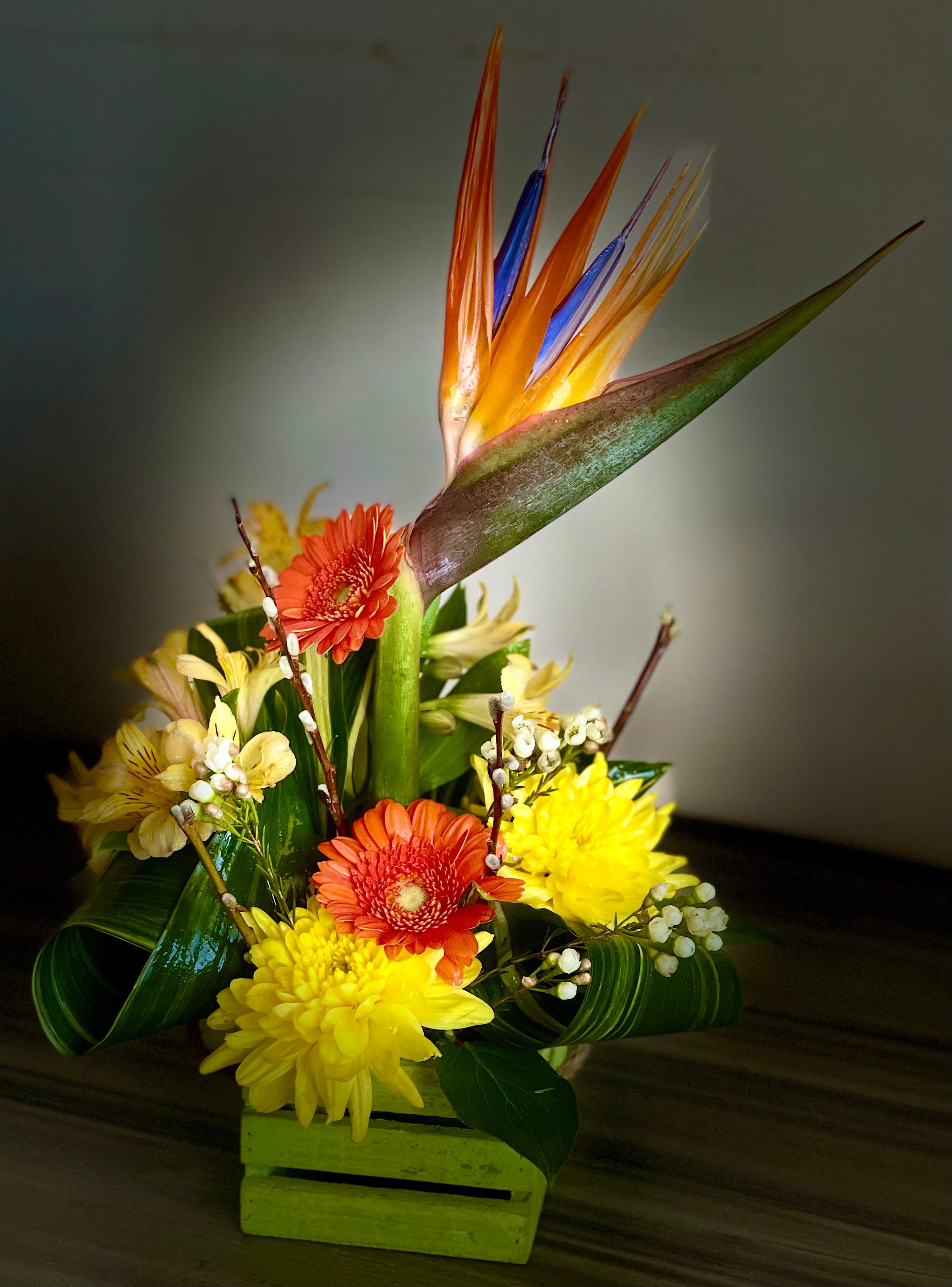 Summer in Style arrangement with a bird of paradise, orange gerbera daisies, and yellow mums in a small green wooden crate
