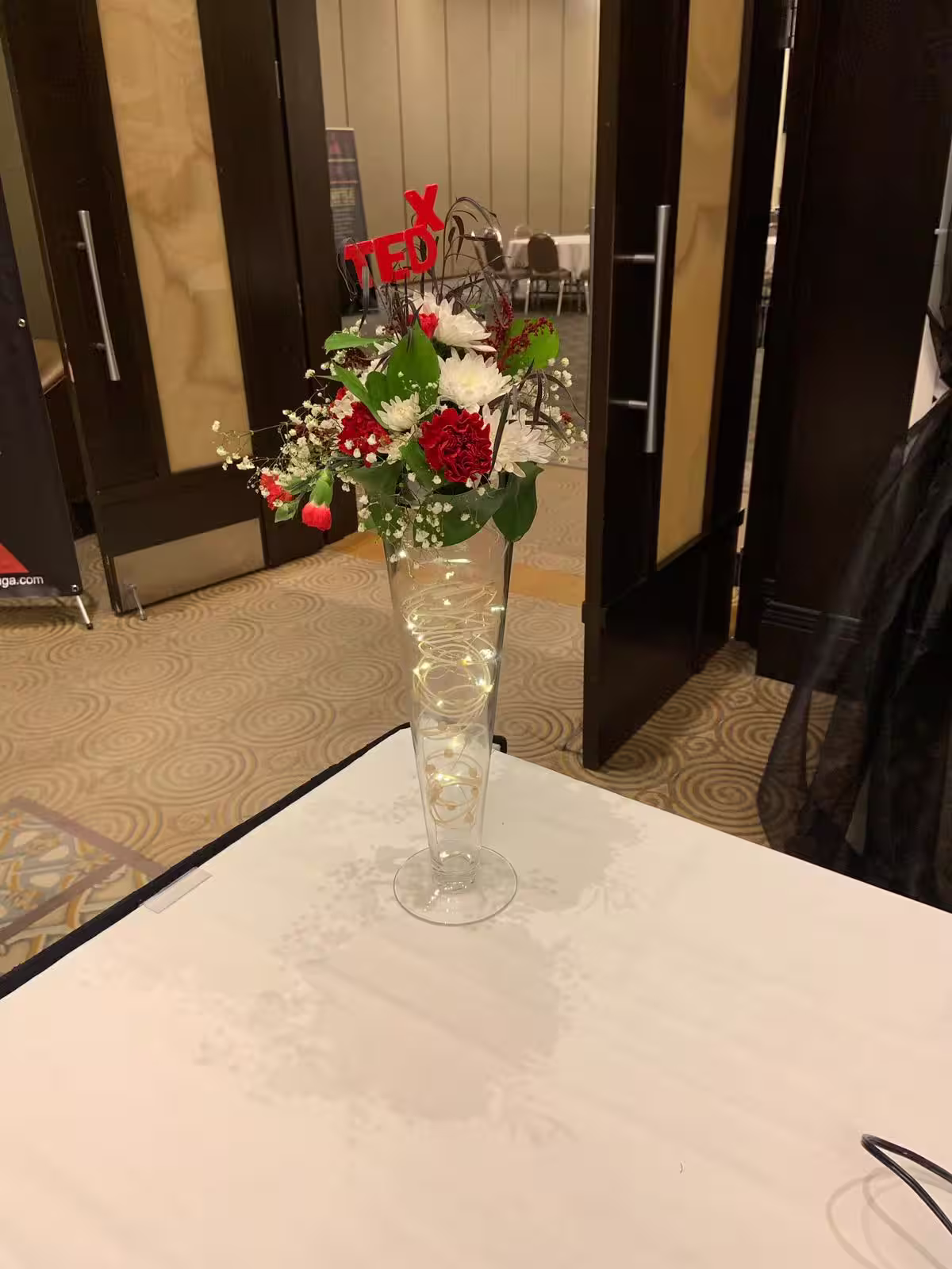 A red and white floral arrangement with fairy lights in a tall glass vase, staged near rows of seating for a speaker event