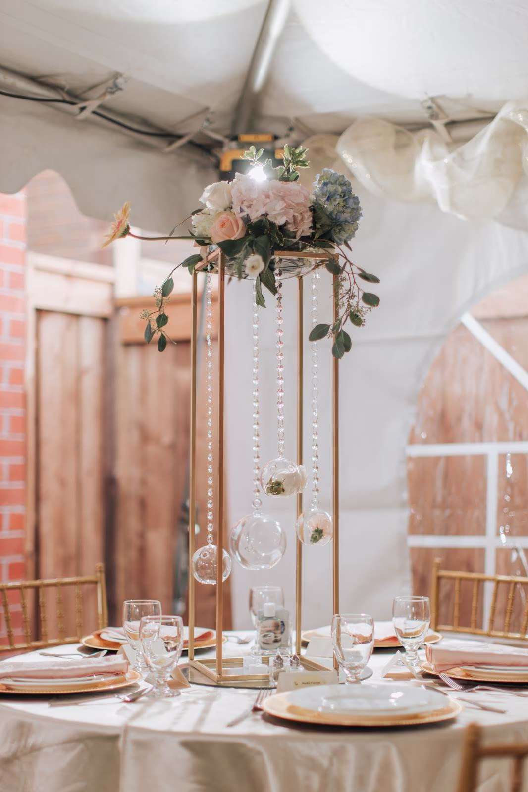 Tall gold stand centrepiece with blush roses, blue hydrangea, and hanging glass votives at a tented reception