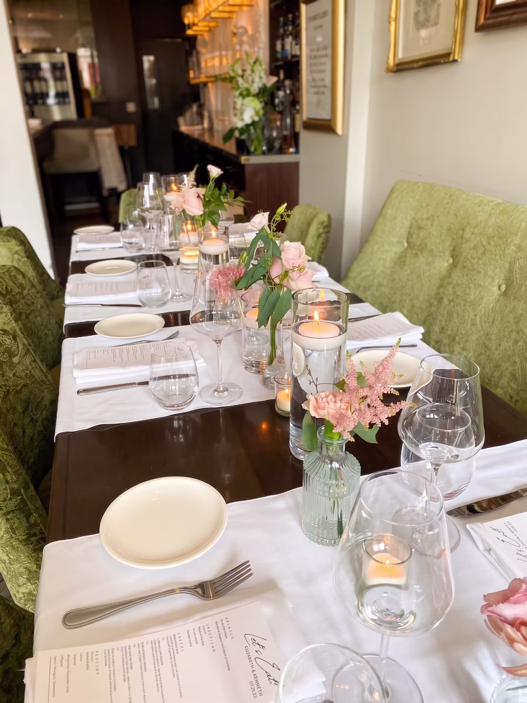 Pink flowers in bud vases with glass candle holders and white place settings along a reception table.